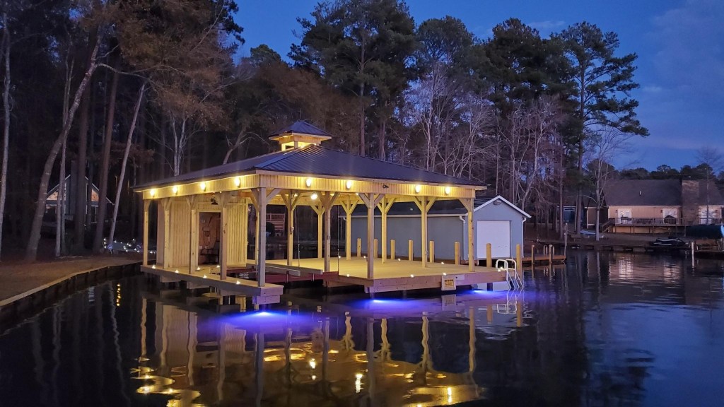 A beautifully illuminated wooden dock at twilight, featuring a metal roof, surrounded by water and trees.